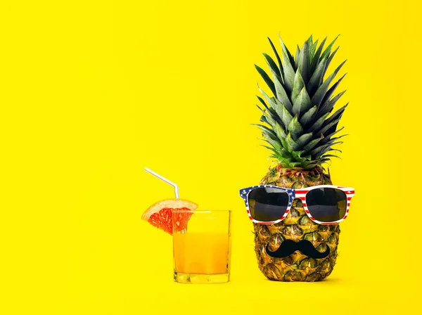 Semester och resor koncept. Roligt mode ananas med Amerika Usa stil flagga stil solglasögon och mustasch med färsk tropisk cocktail med frukt över gul bakgrund. — Stockfoto