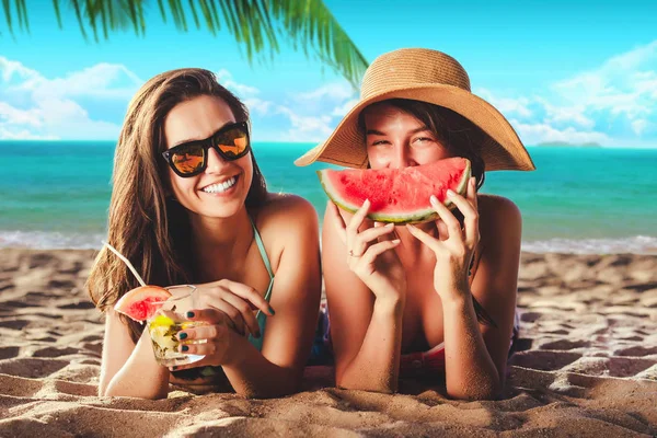 Vacation. Summer travel. Two girls young women in bikinis sunbathing on the tropical beach with cocktails and watermelon in sunglasses smiling at camera laying on the sand with palm and sky background — Stock Photo, Image