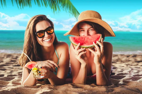 Tatil. Yaz seyahat. İki kız genç kadınlar bikinili kokteyller ve karpuz palmiye ve gökyüzü arka plan ile kum üzerinde döşeme kamera gülümseyen güneş gözlüğü ile tropik Sahilde güneşlenmek