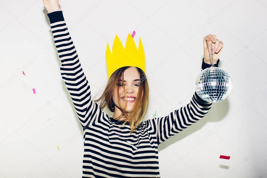 Birthday party, new year carnival. Young smiling woman on white background celebrating brightful event, wears stripped dress and yellow crown. Sparkling confetti, having fun, dancing, laugh, smile.
