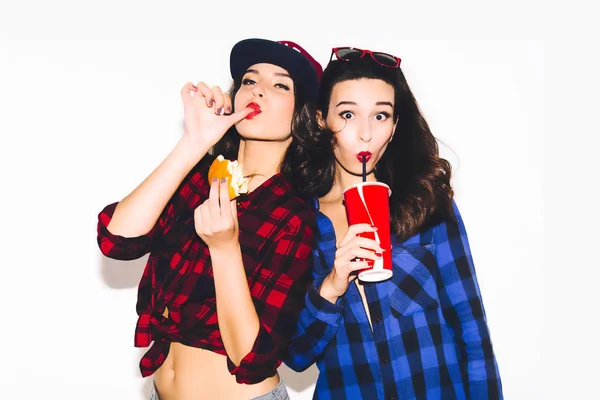 Jóvenes chicas hipster divirtiéndose bebiendo un refresco de paja y sosteniendo una hamburguesa, sonrisa feliz y risa en el fondo blanco . —  Fotos de Stock