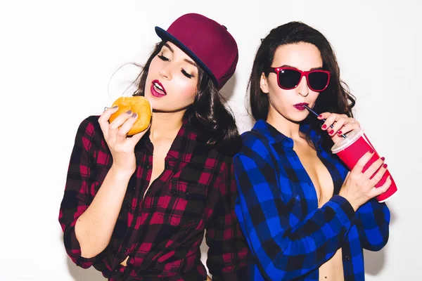 Jovens hipster meninas se divertindo bebendo um refrigerante de palha e segurando um hambúrguer, sorriso feliz e rir no fundo branco . — Fotografia de Stock