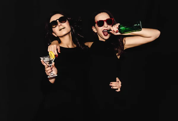 Duas mulheres bonitas vestido de moda noite preto posando isolado em um fundo preto. Menina morena bonita amigos gêmeos se divertindo bebendo coquetéis. Cantando e dançando. Copo de garrafa de álcool . — Fotografia de Stock