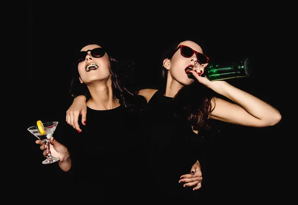 Две красивые женщины черная ночь платье моды позировать изолированы на черном фоне. Симпатичные брюнетки подружки близнецы веселятся, выпивая коктейли. Пение и танцы. Бутылочный стакан алкоголя . — стоковое фото