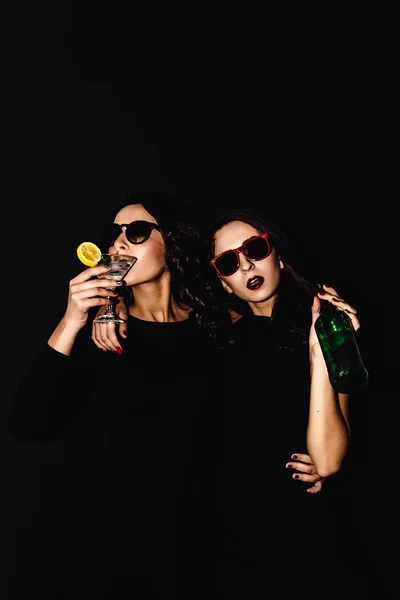 Due belle donne abito nero notte moda posa isolata su uno sfondo nero. Bella bruna ragazza amici gemelli divertirsi bere cocktail. Cantando e ballando. Bottiglia di vetro di alcool . — Foto Stock