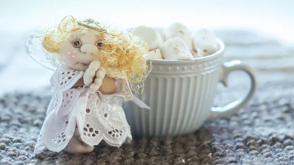 angel in white lace dress with hands folded in prayer. homemade sock toy with fluffy red hair in background of window. cup of marshmallows on knitted napkin
