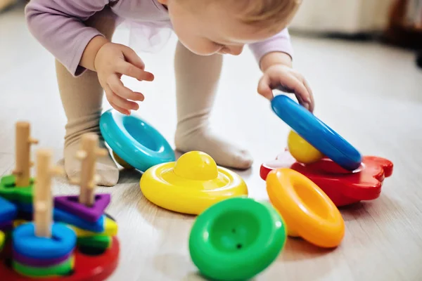 girl lifts pyramid rings from white pod in living room. Montessori wooden toy folded pyramid. Multi-colored toy blue, yellow, red, green.