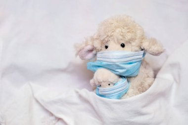 Küçük oyuncak koyun annesi elinde kuzu, elinde fonendoskopla yatakta yatıyor. Çocuk oyunlarında koruyucu tıbbi maske takmak. Koronovirüs, karantina, salgın, salgın, grip, soğuk algınlığı, hastalık. Tıp konsepti ve sağlık