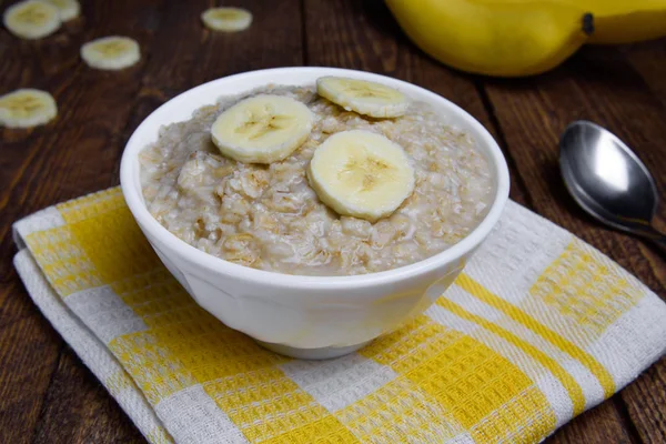 Farina d'avena in una bella ciotola bianca su uno sfondo di legno caldo con fette di banane — Foto Stock