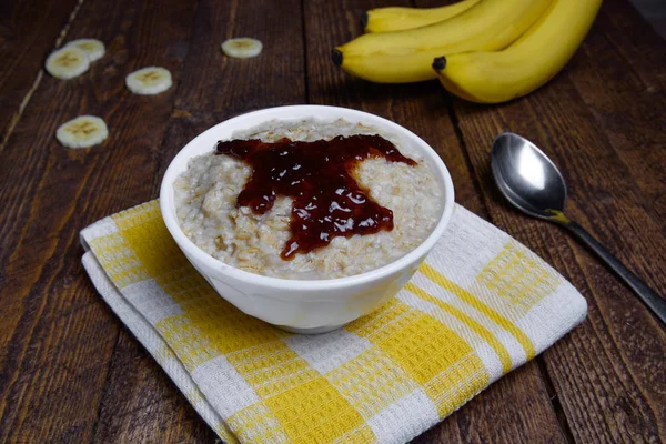 Farina d'avena in una bella ciotola bianca su uno sfondo di legno caldo con fette di banane e marmellata — Foto Stock