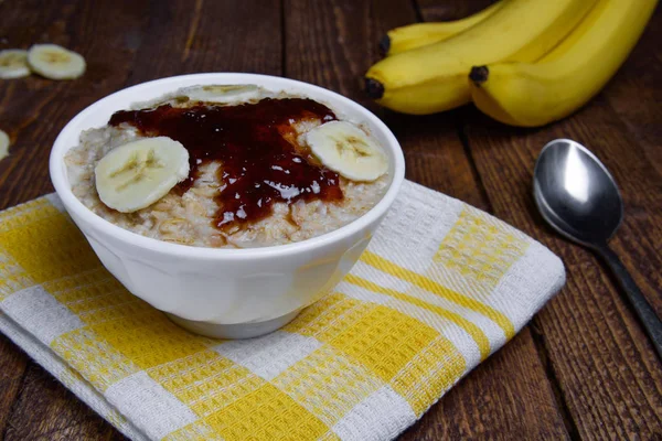 Haferflocken in einer schönen weißen Schüssel auf warmem Holzgrund mit Bananenscheiben und Marmelade — Stockfoto