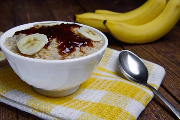 Haferflocken in einer schönen weißen Schüssel auf warmem Holzgrund mit Bananenscheiben und Marmelade — Stockfoto