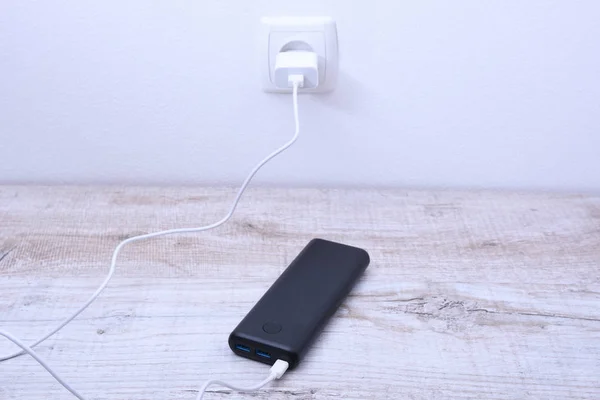 Black phone power bank plugged in the socket on the wall for charging on wooden background