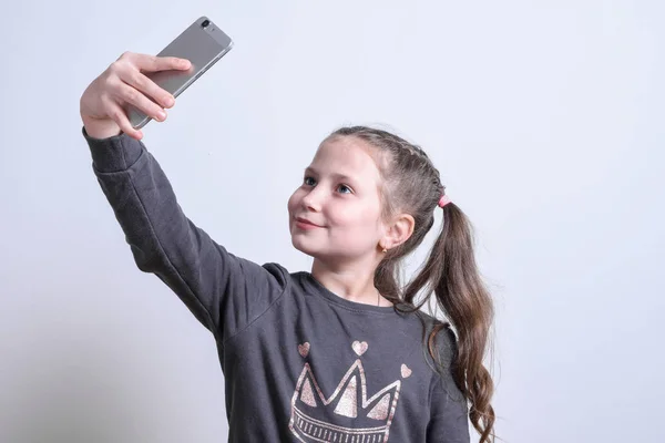 Porträtt av en liten vit flicka gör selfie på en mobiltelefon på en grå bakgrund — Stockfoto