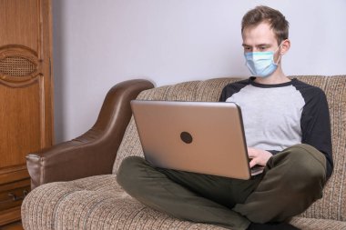 Maskeli genç adam evde, kanepede dizüstü bilgisayar kullanarak çalışıyor. Karantina, kendini izole etme, koronavirüs koruması. Tatil.