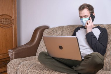Maskeli genç adam evde, kanepede dizüstü bilgisayar kullanarak çalışıyor. Karantina, kendini izole etme, koronavirüs koruması. Tatil.