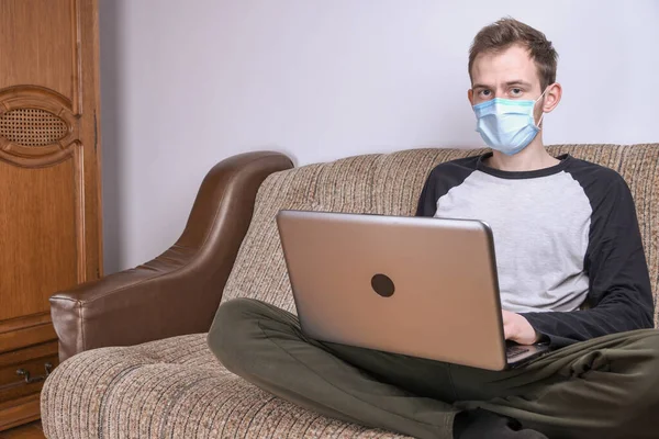 Maskeli genç adam evde, kanepede dizüstü bilgisayar kullanarak çalışıyor. Karantina, kendini izole etme, koronavirüs koruması. Tatil.