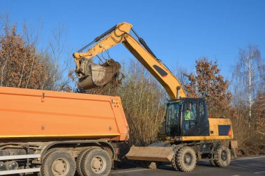 Bir kova kazıcı yol kenarını temizliyor. Yol çalışıyor. Yeni bir yol açıyorum. Kazı kil ve bir taş yükleniyor