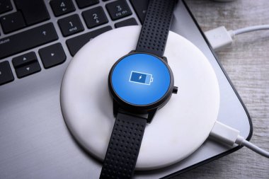 Smart watch on wireless charging with on-screen charging indicator. At the desktop, near at the laptop. Top view. Place for text clipart