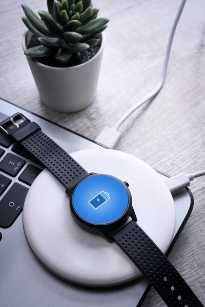 Smart watch on wireless charging with on-screen charging indicator. At the desktop, near at the laptop. Top view. Place for text