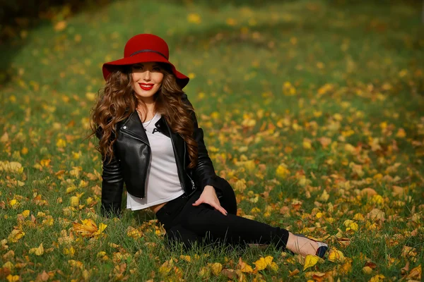 Schöne Frau mit rotem Hut sitzt im Herbst auf dem Rasen im Park. — Stockfoto