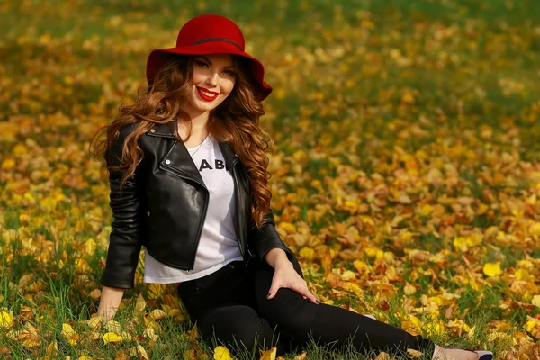 Bella donna in cappello rosso seduta sull'erba nel parco in autunno . — Foto Stock