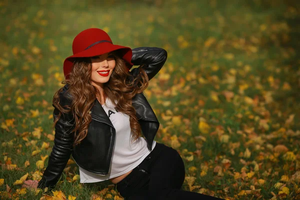 Красивая женщина в красной шляпе сидит на траве в парке осенью . — стоковое фото