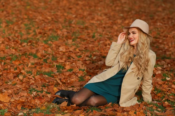 Junge Frau lächelnd im Herbst auf dem Rasen sitzend. Herbst gelben Ahorn Garten Hintergrund. schöne Mädchen in Mantel und Schlapphut im Freien an einem schönen Herbsttag. — Stockfoto