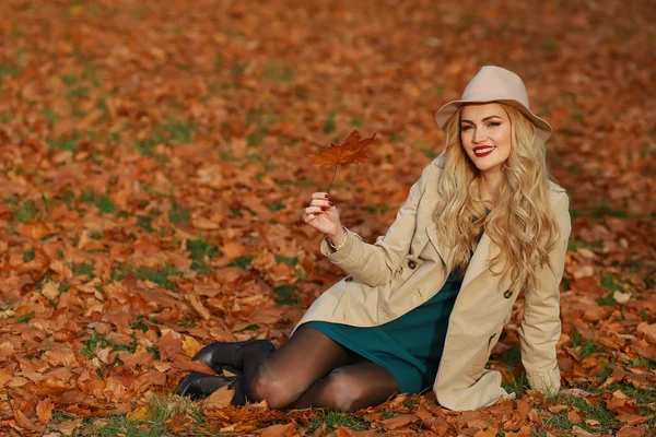 Young woman smiling sitting on the grass in the autumn. fall yellow maple garden background. Beautiful girl in coat and floppy hat outdoors on beautiful fall day. — Stock Photo, Image