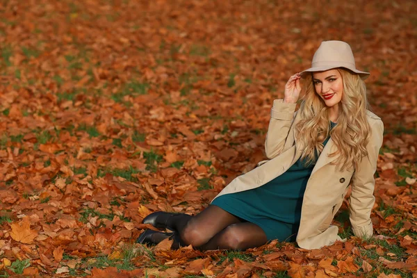 Młoda kobieta uśmiechając się, siedząc na trawie, na jesieni. upadek tle ogród żółty klon. Piękna dziewczyna w płaszcz i miękki kapelusz na zewnątrz na dzień pięknej jesieni. — Zdjęcie stockowe