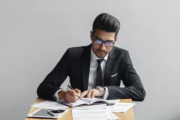 Geschäftsmann schreibt in einem Notizbuch auf grauem Hintergrund in einem Café, Tablet, Telefon und Papiere auf dem Tisch — Stockfoto