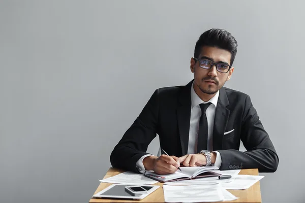 Geschäftsmann schreibt in ein Notizbuch und blickt in die Kamera auf grauem Hintergrund, Tablet, Telefon und Papiere auf dem Tisch. Kopierraum — Stockfoto