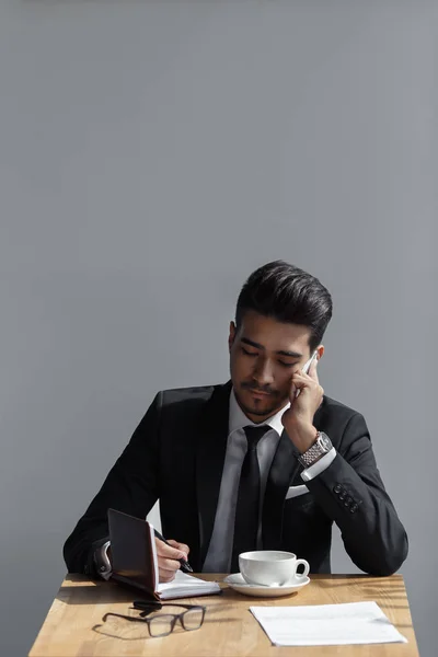 Um belo homem de sucesso falando no escritório de telefone e escrevendo em um caderno no café — Fotografia de Stock