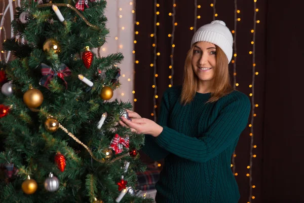 Bella ragazza in un berretto e un maglione decora un albero di Natale — Foto Stock