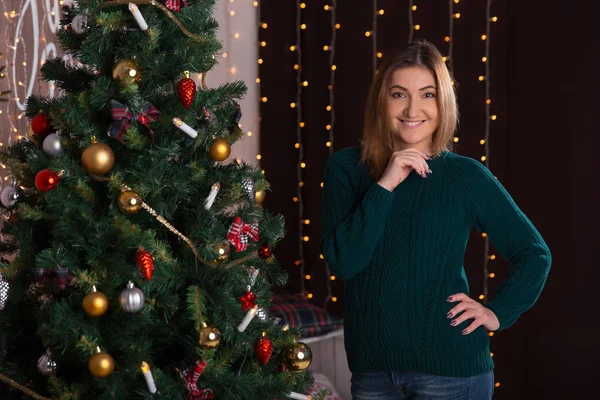 Gyönyörű fiatal nő, mosolygós és a karácsonyfa közelében áll. — Stock Fotó