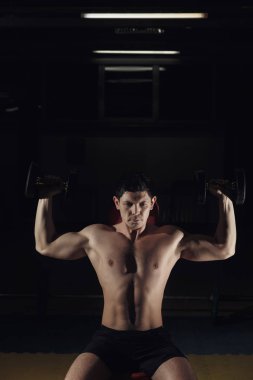 vücut geliştirmeci adam onun elinde iki halter vardır, spor salonunda bir bankta oturuyor.