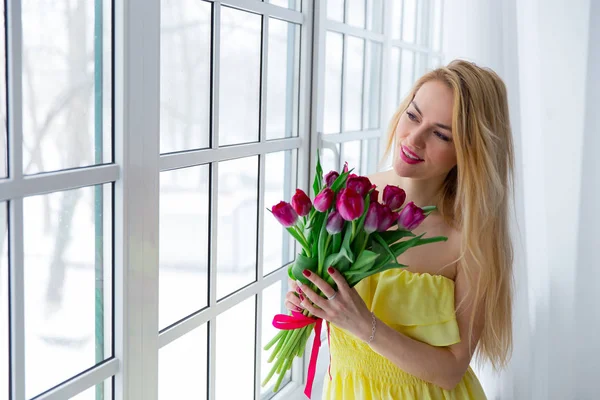 Young beautiful woman with tulip bunch in yellow dress looks at window.