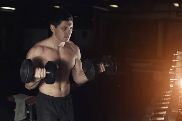 Мускулистый человек тренируется в спортзале, делая упражнения с гантелями — стоковое фото