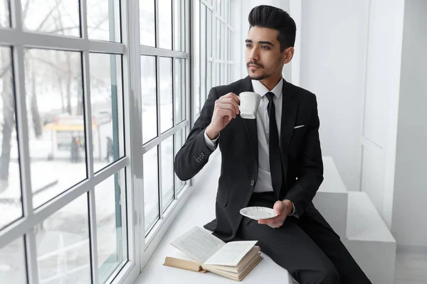 Pensando hombre de negocios en traje sentado en el alféizar de la ventana y bebe café — Foto de Stock