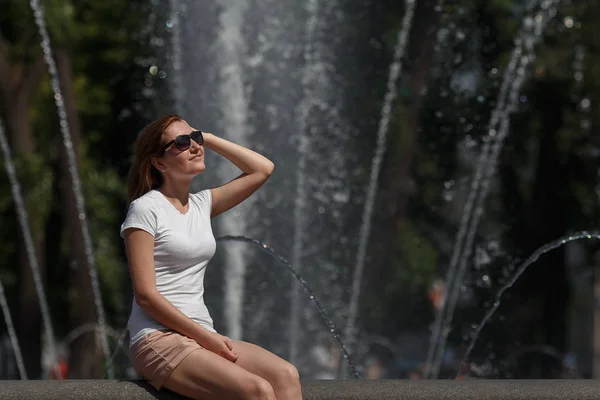 Hübsches junges Mädchen sitzt mit Sonnenbrille auf dem Brunnenhintergrund — Stockfoto