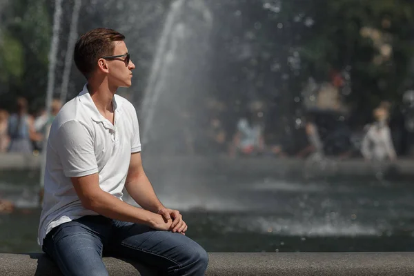 Hombre guapo sentado en gafas de sol en el fondo de la fuente, lejos de la cámara . — Foto de Stock