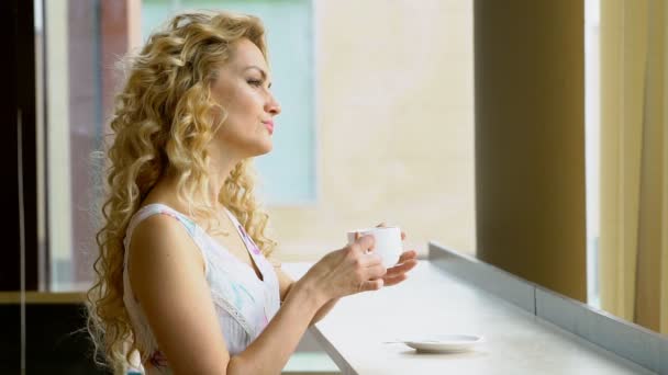 Gyönyörű szőke lány iszik egy csésze forró kávé vagy tea a kávézóban — Stock videók