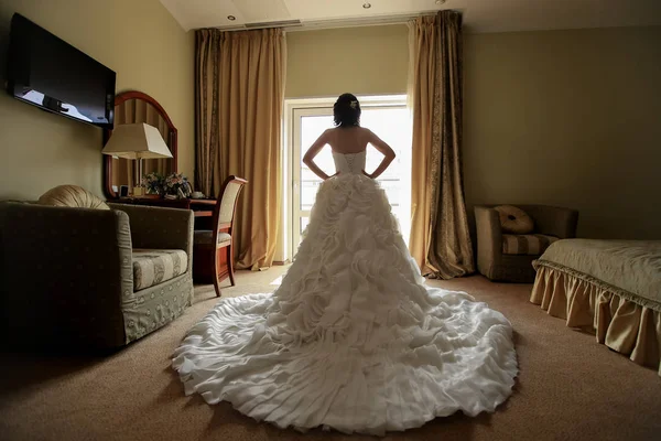 Νύφη σε ένα λευκό φόρεμα με μια μακρά τρένο στέκεται στο παράθυρο στο ξενοδοχείο — Φωτογραφία Αρχείου