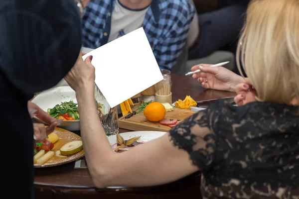 Personas cenando juntas mientras están sentadas en la mesa de madera. Mano femenina sosteniendo papel blanco — Foto de Stock