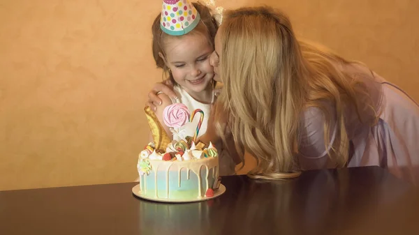 Madre besando a su hija en la fiesta de cumpleaños. pastel de cumpleaños en la mesa —  Fotos de Stock