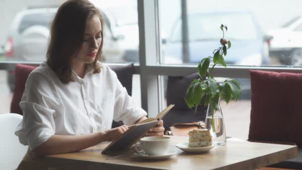 Το κορίτσι διαβάζει ένα βιβλίο κάθεται σε μια καφετέρια και απολαμβάνοντας καφέ και ένα κέικ — Αρχείο Βίντεο