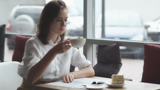 Ελκυστική νεαρή γυναίκα αφιερώνει ελεύθερο χρόνο κάθεται σε καφετέριες, απολαμβάνοντας τον φρέσκο καφέ και διαβάζοντας ένα περιοδικό των γυναικών — Αρχείο Βίντεο