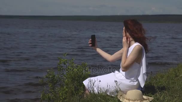 Sensuele close-up portret van mooi meisje in zomer witte jurk op de rivier. meisje maakt een selfie buitenshuis op een smartphone camera — Stockvideo