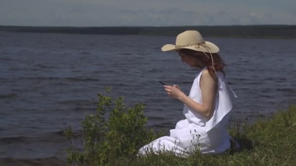 Όμορφη κοκκινομάλλα κοπέλα σε έναν λευκό καλοκαιρινό φόρεμα χρησιμοποιεί ένα κινητό τηλέφωνο που κάθεται στην ακτή και ποτάμια — Αρχείο Βίντεο
