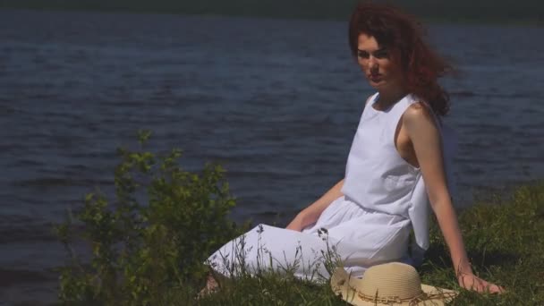 Retrato de una hermosa chica pelirroja en un vestido de verano. mujer joven sentada en la orilla del río y disfrutar del día de verano — Vídeo de stock